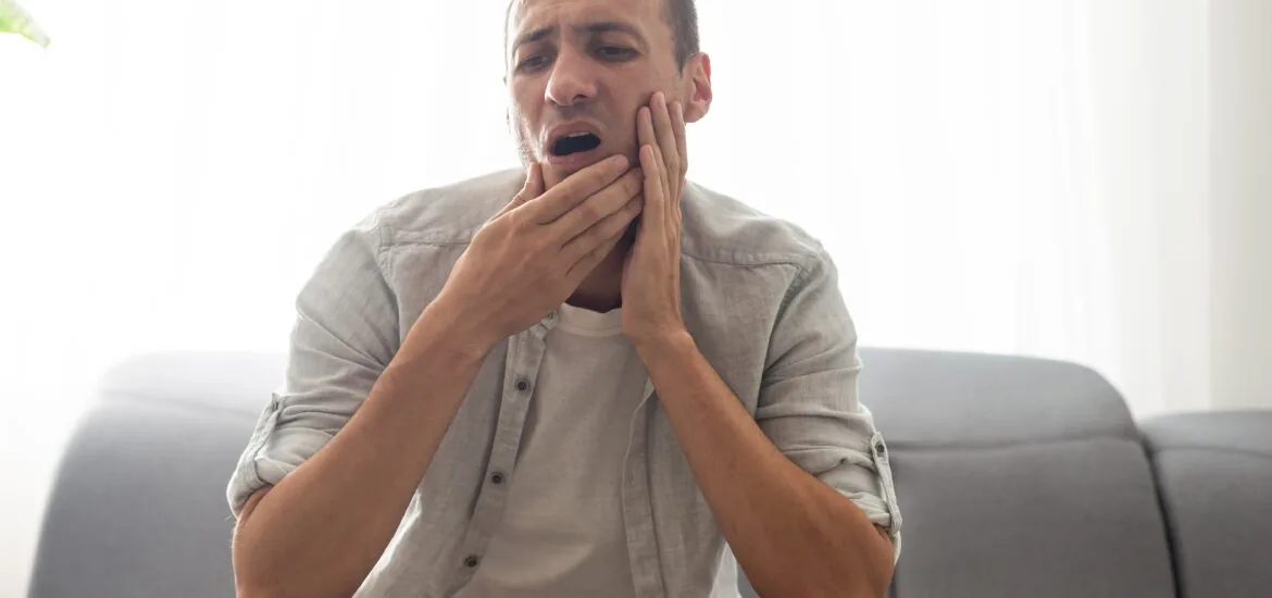 Young man feeling pain, holding his cheek with both hands, suffering from jaw pain